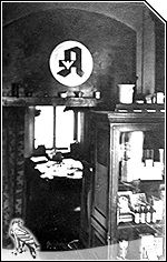 Altes Schwarz-Weiß-Foto. Durchblick zum Büro. Im Vordergrund sehen Sie eine Glasvitrine mit Arzneimitteln.  An der Wand sehen Sie das damals gebräuchliche Apotheken "A".